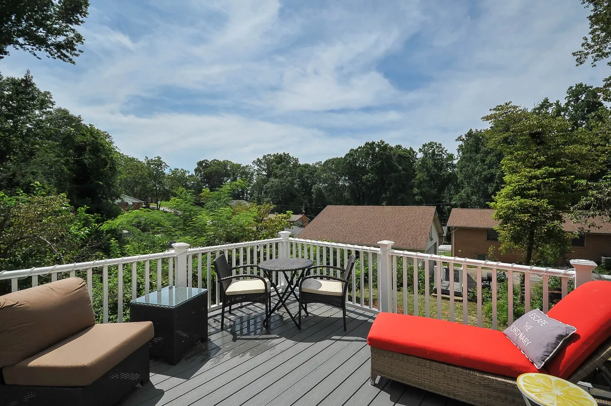 An inviting outdoor deck area featuring a red couch, glass table, black chairs, and wooden railing set against a backdrop of green foliage. The ambiance is cozy and serene, suggesting a perfect place to unwind, relax, or share a quiet evening surrounded by nature.