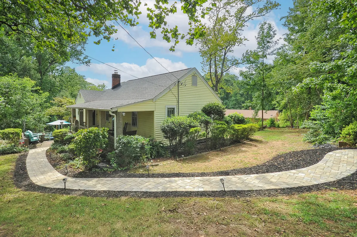 Depicts the exterior of a charming yellow house with a winding stone pathway, framed by lush greenery. The tranquil, welcoming mood suggests a peaceful return after a day out, and the porch hints at sunset viewing and relaxed gatherings.
