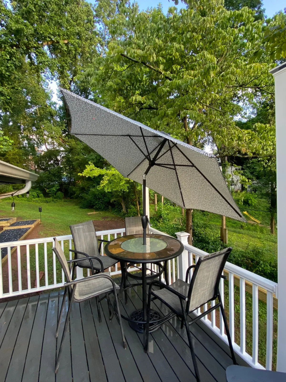 A glass-topped table with black metal chairs sits beneath a large umbrella on a deck, enveloped by vibrant greenery. The scene hints at leisurely outdoor dining or sipping morning coffee while watching the forest come alive, perfectly embodying the idea of seamless indoor-outdoor living.