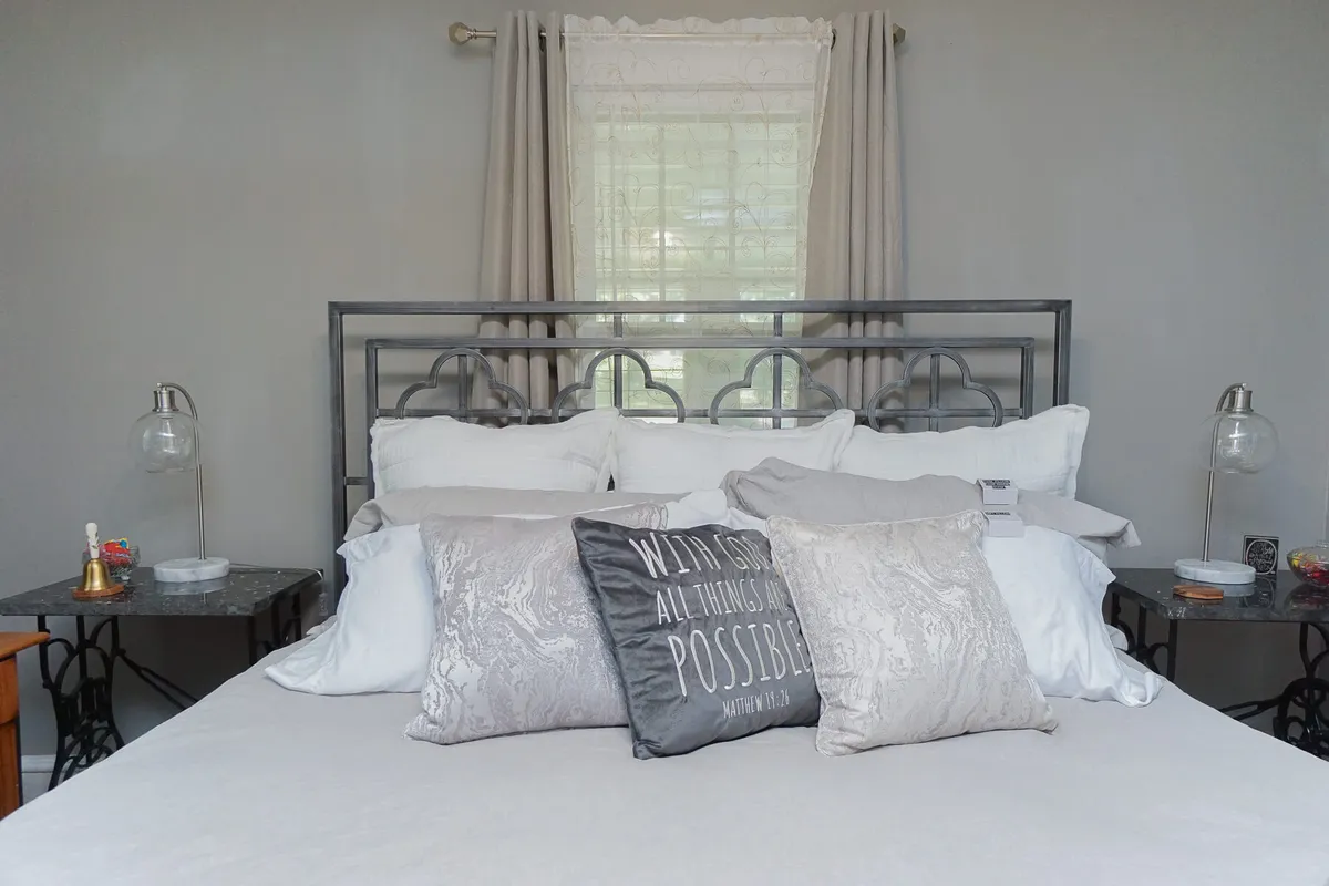 A serene bedroom featuring a neatly made bed with decorative pillows, flanked by nightstands topped with lamps. Soft, sheer curtains filter natural light through the window, creating a peaceful atmosphere ideal for rest and relaxation.