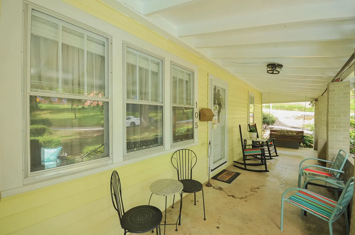 This image captures the yellow exterior of the house with charming rocking chairs and a small table on the front porch, alongside the front door. The scene is bathed in warm light, suggesting either a tranquil morning or a relaxing evening, reinforcing the sense of farmhouse nostalgia and comfort.