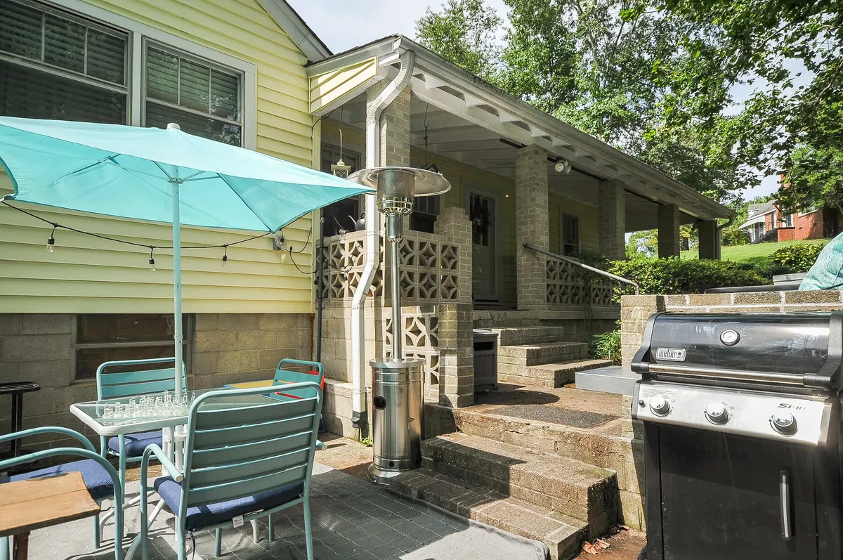 This image showcases a welcoming outdoor dining area equipped with a grill, patio heater, and surrounded by lush greenery. The space is set up for barbecuing, outdoor dining, and socializing under a sunny umbrella, capturing a lively yet relaxed atmosphere ideal for small gatherings or evening get-togethers.