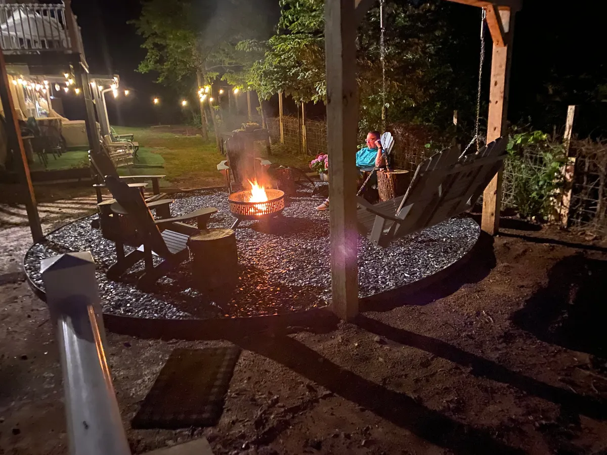 A cozy fire pit area under string lights, complete with a swing and comfortable seating, set amidst lush greenery. The string lights cast a warm glow, creating a magical ambiance ideal for evening gatherings, storytelling, and stargazing.