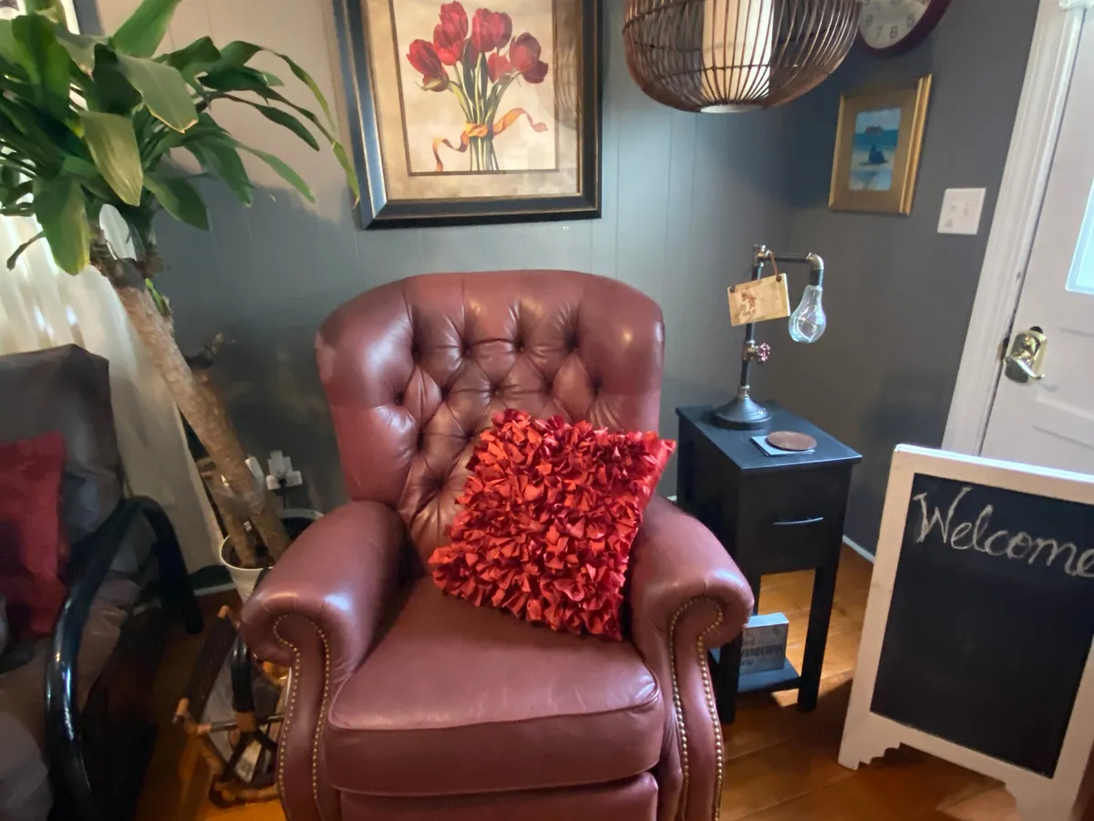 A welcoming corner featuring a plush armchair with a red decorative pillow, framed artwork, and a 'welcome' sign. The setting offers vintage-inspired charm and a cozy spot for relaxing or reading, awash with soft, natural lighting.