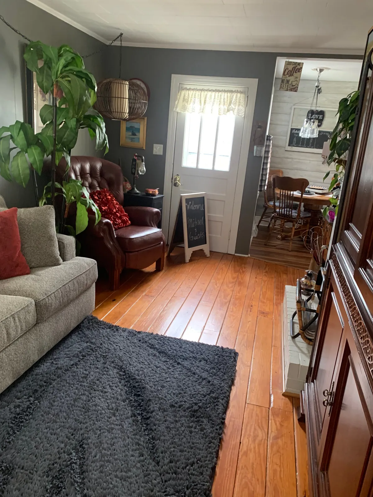 A cozy entryway and living room with a comfortable sofa and armchair, a welcoming rug, a prominent 'welcome' sign, and soft lighting. The décor combines vintage touches with a warm, homey feel. There’s a sense of space for both gathering and relaxing, matching the farmhouse vibe.