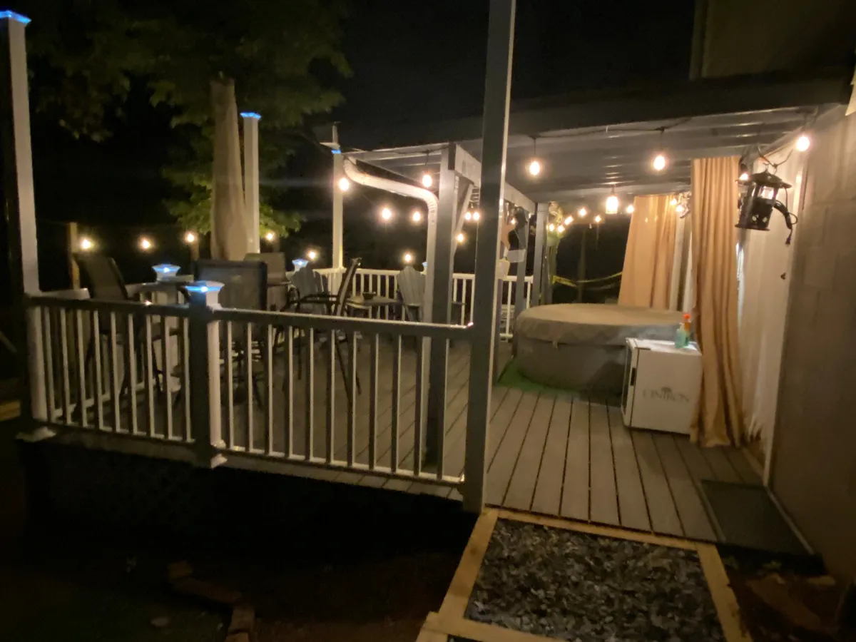 A cozy, inviting backyard scene at dusk features twinkling string lights overhead, a bubbling hot tub, a comfortable outdoor seating area, and a decorative lantern casting a warm glow, all nestled amidst leafy trees. The setting promises intimate relaxation and memorable evenings under the stars.