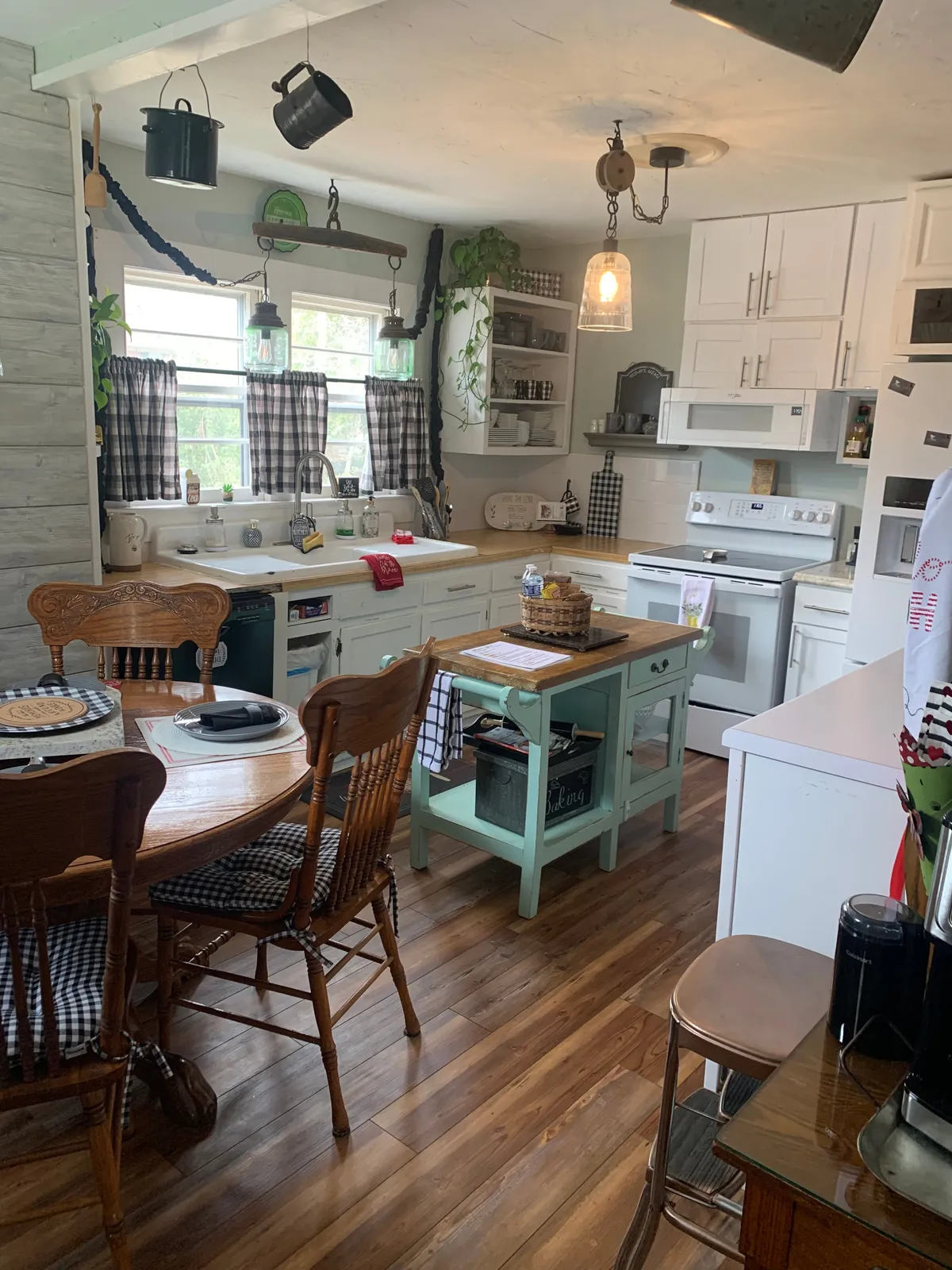 This interior image showcases a kitchen island and dining table positioned near windows with checkered curtains and hanging lights. The room is flooded with natural light, creating a warm, inviting atmosphere ideal for family gatherings, shared meals, and morning coffee rituals. The visible layout makes it easy to picture conversation flowing between the kitchen and dining spaces.